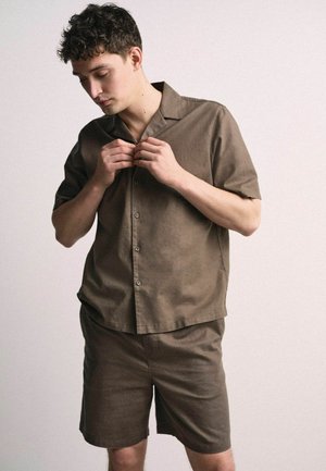 Bărbat tânăr cu păr scurt și creț nasturând o cămașă maro cu mâneci scurte, purtând pantaloni scurți maro asortați, stând în fața unui fundal simplu.