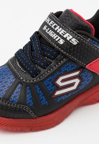 Czarne, niebieskie i czerwone sneakersy Skechers S-Lights z rzepem, wzorzystymi bokami i czerwonym spodem na białym tle.