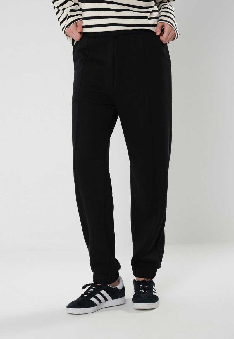 Sorte joggebukser med elastiske mansjetter og sidesømmer, laget av mykt stoff. Kombinert med svart-hvite joggesko og en stripete langermet topp.