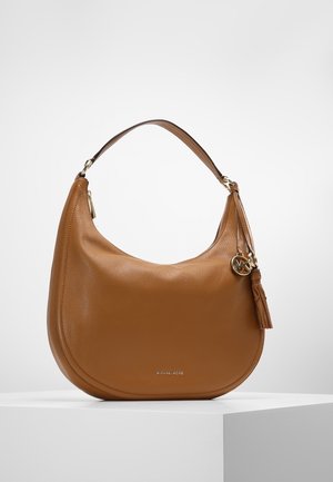 Rdzawy skórzany damska torba na ramię Michael Kors o zakrzywionym kształcie, z zawieszką w postaci złotego logo i ozdobnym frędzlem, ustawiona na białym postumencie.