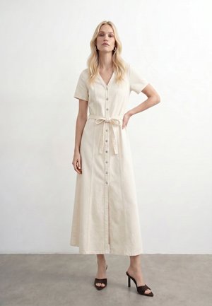 Blondynka w kremowej, zapinanej na guziki sukience midi z krótkimi rękawami i przewiązanym paskiem w talii, stojąca na tle gładkiej białej ściany.