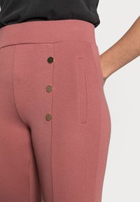 Rosa byxor med texturerad yta, med hög midja och tre knappar i guldton på sidan. Detalj med en enkel sidficka.