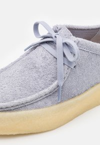 Šedá semišová bota s texturovaným povrchem, kulatou špičkou, nízkým profilem a světle modrou šněrovací stuhou uvázanou do mašle. Béžová gumová podrážka.