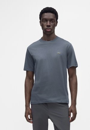 Jeune homme à la peau foncée portant un t-shirt gris uni à manches courtes avec un petit logo vert sur la poitrine et un pantalon gris assorti.