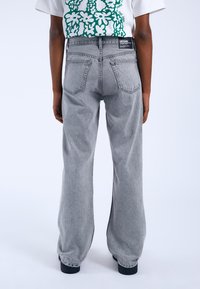 Los pantalones anchos de mezclilla gris presentan una cintura alta, bolsillos traseros y detalles de costura sutiles. La tela tiene un acabado ligeramente texturizado.