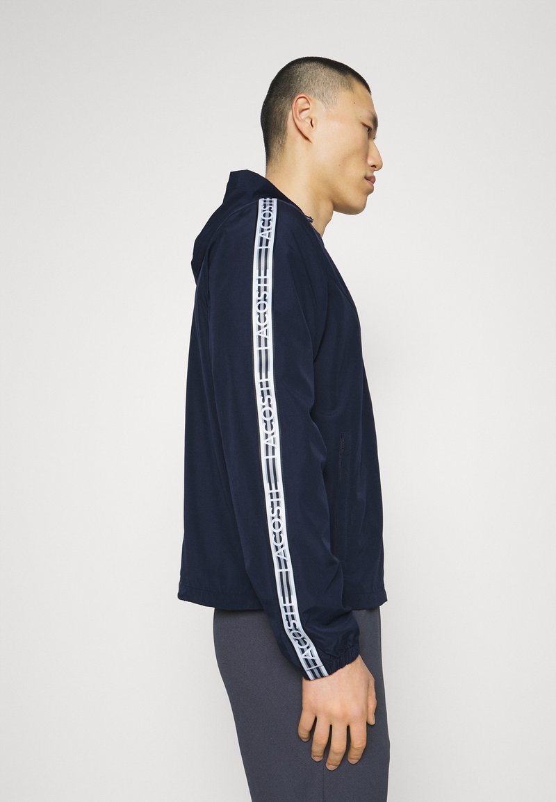 Marineblauer Kapuzenjacke mit kontrastierenden weißen Logostreifen entlang der Ärmel. Verfügt über eine glatte Textur und seitliche Reißverschlusstaschen.