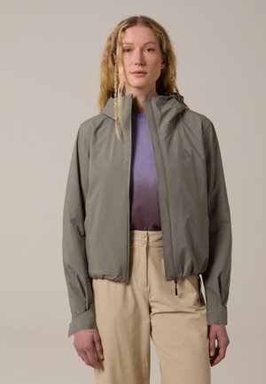 Mujer con cabello rubio largo que lleva una chaqueta gris con capucha sobre una camiseta morada y pantalones beige de cintura alta, de pie contra un fondo liso.