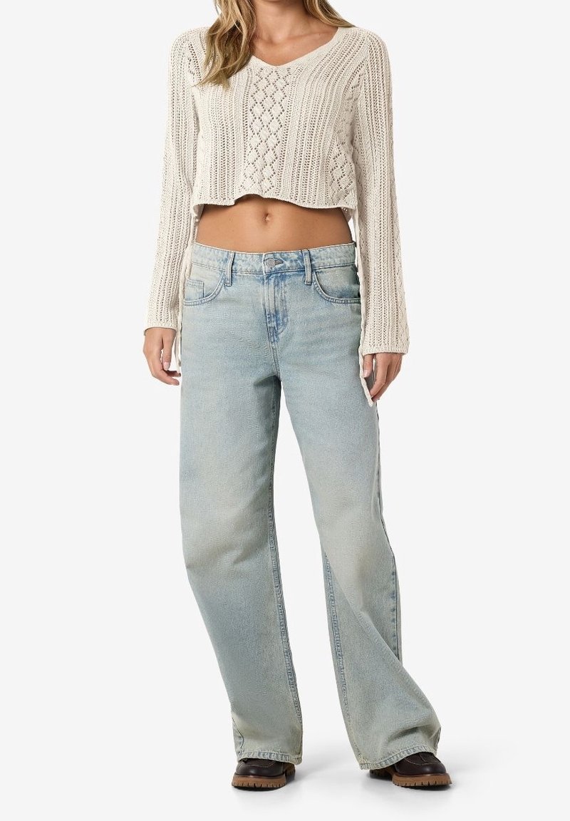 Ljusbeige, kort stickad tröja med texturerad stickning, tillsammans med lösa, ljusblå denimjeans och mörkbruna skor.