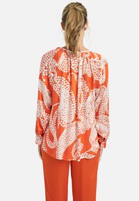 Hedvábná blůza s dlouhým rukávem v živé oranžové barvě s bílým abstraktním vzorem, s nabíraným výstřihem a volně padajícím střihem. Měkká tkanina, uvolněná silueta.