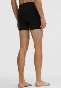 Férfi modell hátulról, fekete, testhez simuló boxeralsót viselve, mezítláb áll egy sima világos háttér előtt.