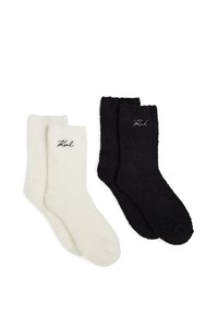 Zwei Paar flauschige Socken; ein weißes, ein schwarzes. Jede Socke hat eine weiche Textur und ist am Bündchen mit schwarzem Stickerei des Markennamens versehen.