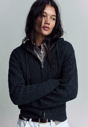 Mlada ženska z dolgimi temnimi lasmi, oblečena v črn pulover z zadrgo in črtasto srajco, zložila roke in gleda v stran.