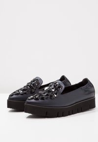 Černé platformové slip-on boty mají hladký svršek s geometrickými černými dekoracemi, elastický límec a texturovanou podešev.