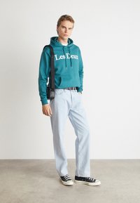 Teal hoodie with "Les Deux" in white, paired with light blue jeans and black sneakers. The model holds a black crossbody bag.