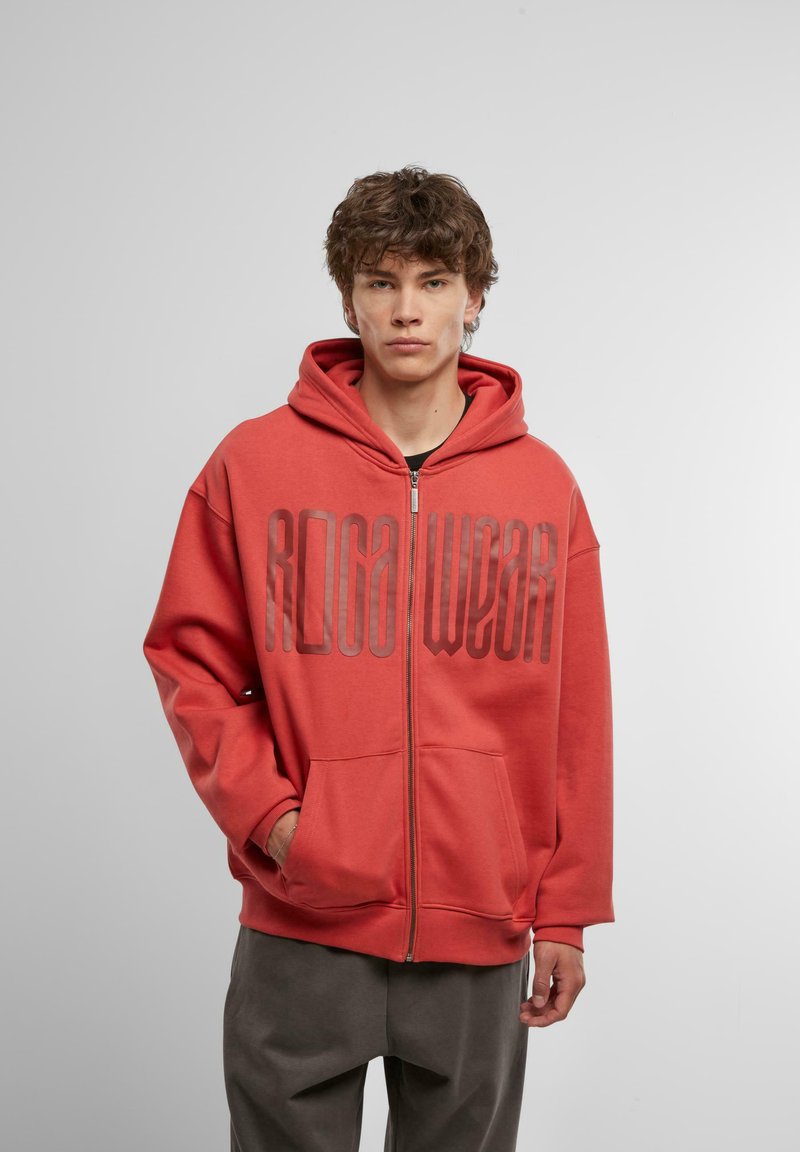 Red zip-up hoodie with a large printed logo "RIGA WEAR" on the front. Soft fabric, relaxed fit, and two front pockets.