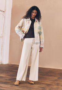 Mehrfarbige, gemusterte, übergroße Jeansjacke mit strukturierten Akzenten, getragen über einem dunklen Oberteil und weiten cremefarbenen Hosen, ergänzt durch hellbraune Schuhe.