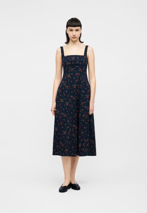 Γυναίκα με ίσια σκούρα μαλλιά που φοράει αμάνικο, μεσαίου μήκους φόρεμα με μπλε ναυτικό χρώμα και λουλούδια, καθώς και μαύρες μπαλαρίνες, σε απλό λευκό φόντο.