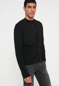 Mustast kootud kardigan V-kaelusega, millel on viis nööpi ja soonilised varrukaotsad, kombineeritud tumedate hallide teksadega.