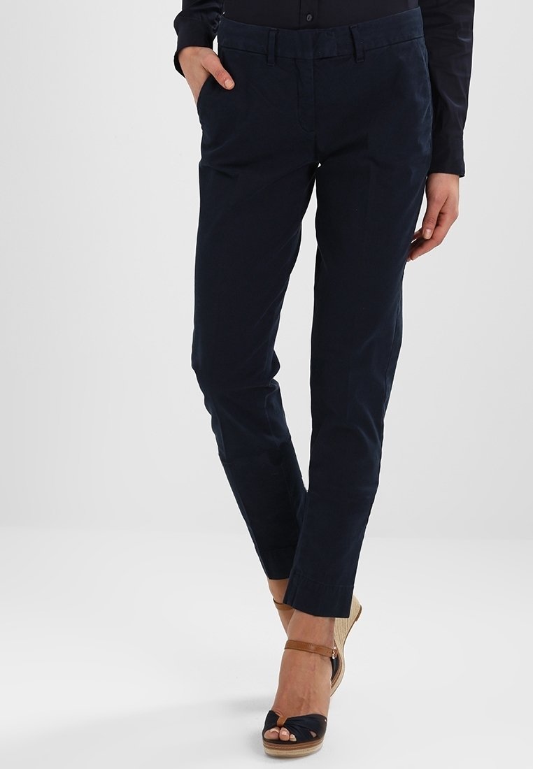 Femme portant un pantalon ajusté bleu marine foncé et des sandales compensées noires avec des brides à la cheville, debout avec une main dans la poche sur un fond blanc.