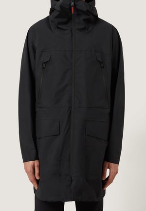 Veste de pluie longue noire avec capuche, zip frontal, deux poches poitrine zippées et deux grandes poches à rabat près des hanches, portée par une personne.