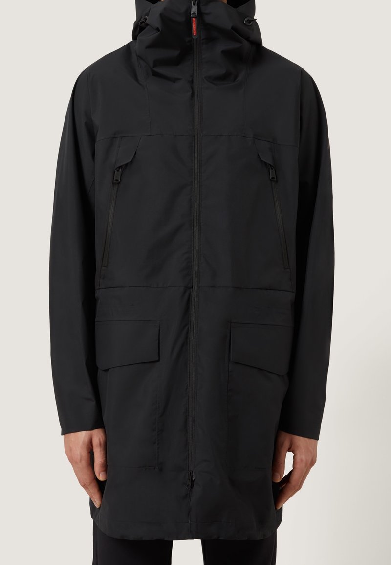Veste de pluie longue noire avec capuche, zip frontal, deux poches poitrine zippées et deux grandes poches à rabat près des hanches, portée par une personne.