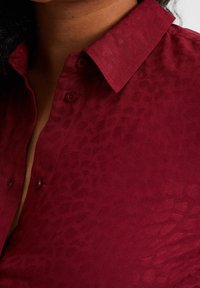 Burgundy knäppskjorta med ett texturerat djurmönster, med klassisk krage och två knappar på knäppningen.