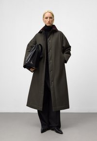 Lungo cappotto verde oliva con colletto scuro, dotato di chiusure a scatto e tasche laterali. Abbinato a pantaloni neri a vita alta e scarpe a punta.