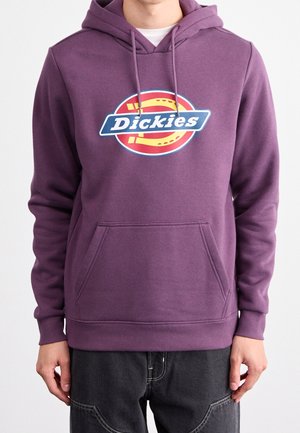 Mann trägt ein lilafarbenes Dickies-Hoodie mit Fronttasche und Kordelzügen, kombiniert mit einer schwarzen Hose mit sichtbaren Nähten.