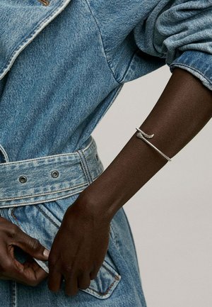 Pulsera de serpiente de plata sobre un brazo oscuro, llevada sobre una chaqueta de mezclilla azul claro con cintura entallada y tela texturizada. Botones y costuras detallados.