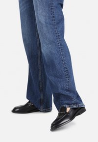 Černé kožené loafers s kulatou špičkou a řetízkovým akcentem, v kombinaci s světle modrými džíny. Boty mají nízký stacked podpatek.
