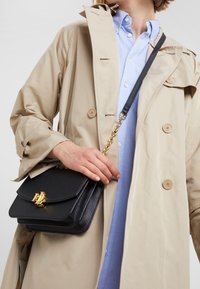 Czarna skórzana torebka z złotymi okuciami, klapą i łańcuszkowym paskiem. Noszona z beżowym płaszczem trencz na jasnoniebieskiej koszuli.