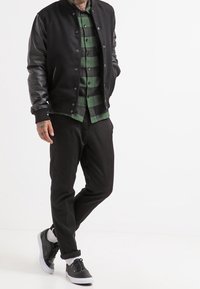 Svart varsityjacka med läderärm över en grön rutig skjorta, kombinerad med svarta byxor och svarta sneakers med kontrasterande vita sulor.