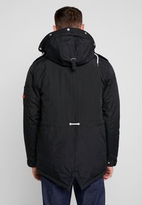 Svart parka-jacka med dragsko i midjan, stor huva och tryckknappar. Har en texturerad utsida och en distinkt böjd fåll.