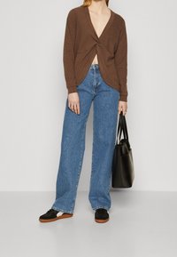 Top marrone a maniche lunghe con dettaglio a nodo in vita, abbinato a jeans larghi blu chiaro e sneakers nere. Porta una borsa tote nera.