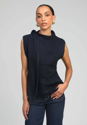 Femme aux cheveux tirés en arrière portant un haut tricoté sans manches de couleur sombre avec un détail écharpe et un jean foncé, posant devant un fond gris uni.