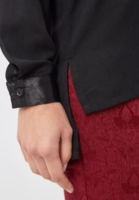 Blusa negra con puños y detalles de botón negros y brillantes, combinada con tejido burdeos texturizado. Línea de costura clara en el lado.