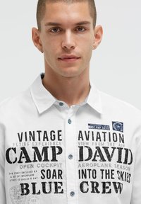 Weiße Hemd mit Knöpfen, das bedruckten schwarzen Text und grafikbezogene Motive zur Luftfahrt zeigt. Blaue Knöpfe und ein Patches akzentuieren den Kragenbereich.