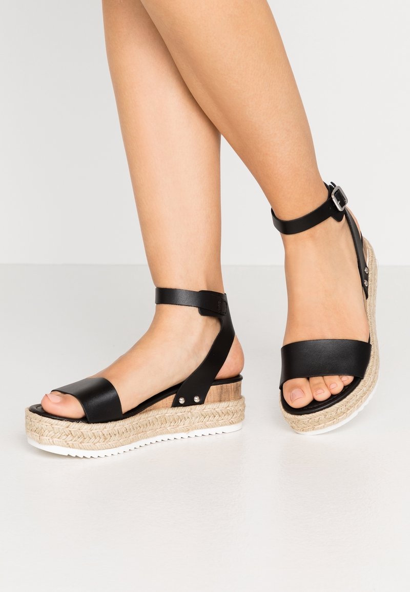 Sandales compensées en cuir noir avec une semelle en jute, dotées d'une bride à la cheville et d'un design à bout ouvert, semelle blanche contrastante et accents argentés.