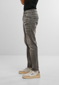 Jeans grii, deteriorate, cu rupturi vizibile și croi slim, stilizate cu adidași bej. Materialul pare a fi denim cu o textură uzată.