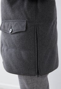 Abrigo de lana gris con un gran bolsillo abotonado, detalle de cremallera lateral y forma estructurada. Se lleva sobre unos pantalones grises con estampado.