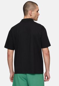 Man med kort hår som bär en enkel svart kortärmad polo och gröna byxor, med ryggen mot en vit bakgrund.