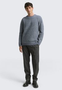 Grauer Strickpullover mit strukturiertem Muster, Rundhalsausschnitt, gerippten Bündchen, kombiniert mit dunkelgrauer Hose und schwarzen Stiefeletten.
