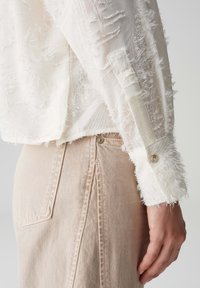 Top bianco a maniche lunghe con tessuto strutturato e dettagli sfrangiati, polsino con bottoni, abbinato a jeans beige chiaro a vita alta.