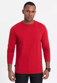 Röd långärmad t-shirt i bomull, med rund hals och åtsittande ärmar, matchad med mörka jeans. Inkluderar en liten logotagg i botten.