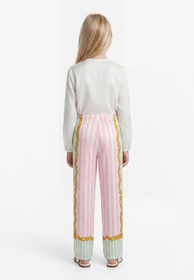 Dieťa s dlhými blond vlasmi stojí chrbtom, má na sebe biely dlhý rukáv a pastelovo pruhované nohavice so zlatými ozdobnými lemami.