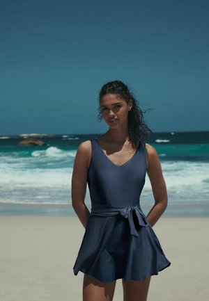 Kvinna med lockigt hår i en ärmlös mörkblå klänning med knuten midja står på en sandig strand med vågor i bakgrunden.