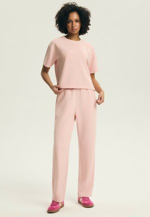 Kobieta o kręconych włosach, ubrana w bladoróżową bluzkę z krótkim rękawem i pasujące szerokie spodnie, do tego jasnoróżowe trampki.