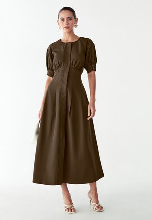 Brun midi-kjole med puffede ærmer og knaplukning foran. Glat tekstur, indsnøret talje og udvidet nederdel. Kombineret med cremefarvede hæle.