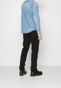 Ljusblå denimskjorta med rundad nederkant och knäppning på ärmarna; kombinerad med svarta slim-fit jeans och svarta snörstövlar, visad från baksidan.