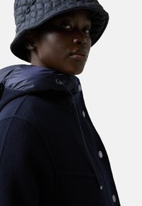 Granatowa kurtka z kapturem, z pikowanym, błyszczącym kołnierzem, z metalowymi guzikiem i sznurkiem do regulacji. Noszona z pasującą kapeluszem typu bucket.